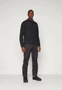 Cămașă neagră cu nasturi, cu mâneci lungi, două buzunare pe piept, asortată cu pantaloni gri închis și adidași negri. Textură netedă, design simplu.