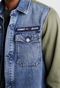 Farkkutakki oliivinvihreillä hihoilla, jossa rintatasku, "TOMMY JEANS" -patch ja nappikiinnitys. Rakenne on tekstuurinen ja värit ovat kontrastiset.