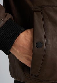 Giacca di pelle marrone con polsini neri a coste, dotata di una tasca laterale e di una toppa circolare del logo sul tessuto. Texture liscia, design aderente.