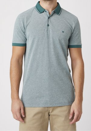 Man wearing a light blue short-sleeve polo shirt with dark green collar and sleeve edges, paired with beige pants.