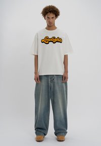 Λευκό φαρδύ μπλουζάκι με την ένδειξη "Lowlights" σε κίτρινα και μαύρα γράμματα, συνδυασμένο με χαλαρές μπλε τζιν παντελόνες και καφέ υποδήματα.