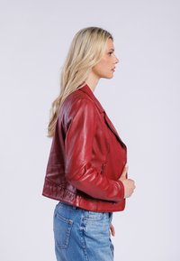 Červená kožená bunda s těsným střihem, upravenými rameny a zipovým zapínáním; v kombinaci se světlými modrými džínami.