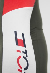 Enganliegendes Sportshirt aus grünem Stoff mit einem auffälligen roten und weißen Grafikdesign auf dem linken Ärmel und Logo-Details.