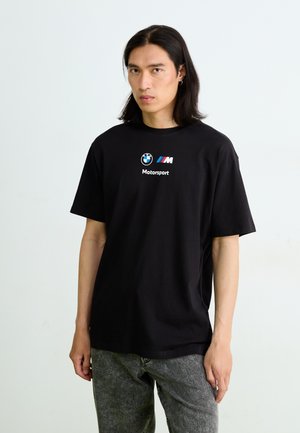 Melnas kokvilnas T-krekls ar BMW logotipu un baltu "Motorsport" tekstu. Īsas piedurknes un brīva piegriezuma, kopā ar pelēcām teksturētām biksēm.