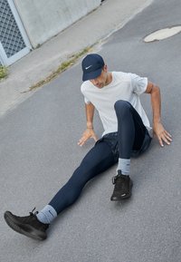 Černé sportovní boty, tmavě modré legíny a světle šedé tričko Nike. Subjekt sedí na asfaltu, má na sobě černou kšiltovku a šedé kotníkové ponožky.