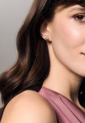 Złote kolczyki wkrętki zaprojektowane w wirującym wzorze, z gładkimi, błyszczącymi powierzchniami. Modelka ma na sobie głęboki fioletowy top bez rękawów.