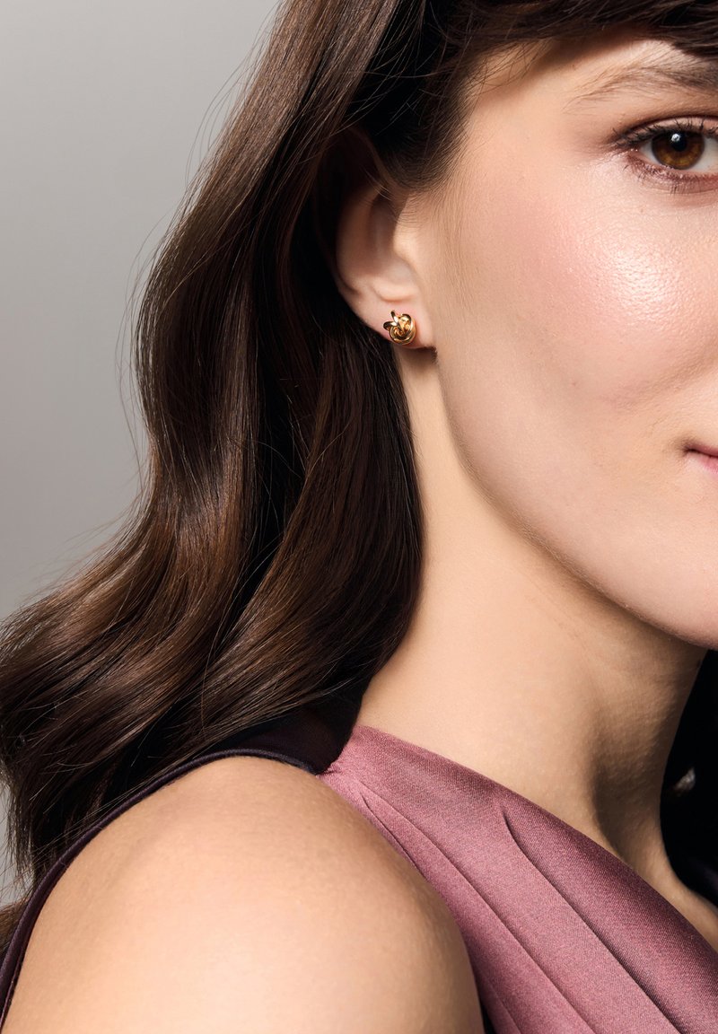 Złote kolczyki wkrętki zaprojektowane w wirującym wzorze, z gładkimi, błyszczącymi powierzchniami. Modelka ma na sobie głęboki fioletowy top bez rękawów.