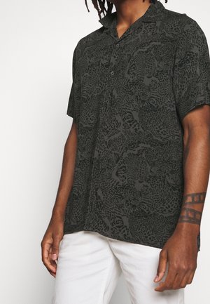 Mann trägt ein dunkles Hemd mit Knöpfen und einem schwarzen Leopardenmuster sowie weiße Hosen, zeigt ein geometrisches Tattoo auf dem linken Unterarm.