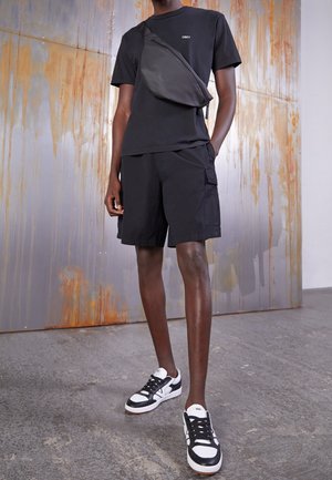 Personne portant un t-shirt noir OBEY, un short cargo noir, un sac bandoulière noir et des baskets Vans noir et blanc, se tenant sur un sol en béton.