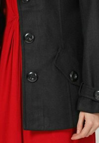 Abrigo de lana negro con un diseño de doble botonadura, que presenta grandes botones negros y un dobladillo curvado. Un vestido rojo es parcialmente visible debajo.