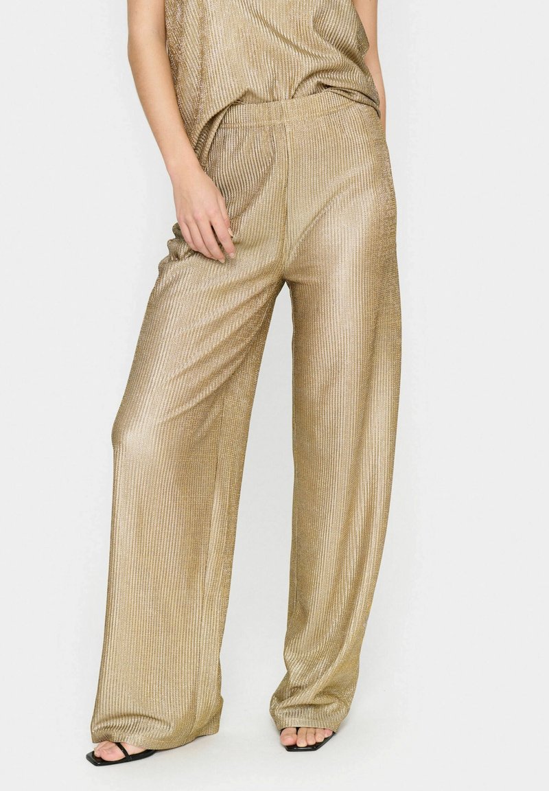 Metaliczne złote spodnie typu wide-leg o żeberkowanej fakturze i elastycznym pasie, w połączeniu z luźno dopasowaną górą w tym samym stylu. Widać płaskie czarne sandały.