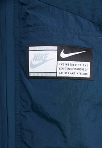 Tumši zila auduma virsma ar gludu tekstūru, kurā ir melns uzlīme ar baltām un gaiši ziliem Nike logo un tekstu "Izstrādāts pēc precīzām specifikācijām."