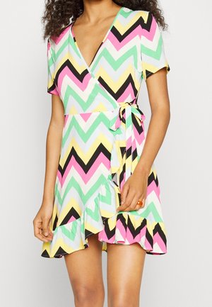 Femme portant une robe portefeuille à manches courtes avec des motifs zigzag pastel en rose, jaune, vert, noir et blanc, nouée à la taille avec un ourlet à volants.