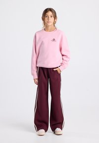 Jeune personne debout portant un sweat-shirt rose avec le logo "Golden Bagel", un pantalon large bordeaux avec des rayures blanches et des baskets de couleur claire.