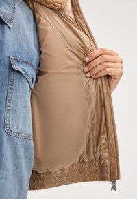 Jasnobrązowa kurtka z miękkim, teksturowanym, jasnobeżowym kołnierzem z futra. Wnętrze ma gładką, błyszczącą beżową podszewkę z elastycznym wykończeniem.