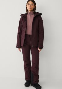 Bordeaux ski jack met een ritssluiting aan de voorkant, bijpassende broek en een lichtpaarse fleece top. Zwarte accenten op de broek en het schoeisel.
