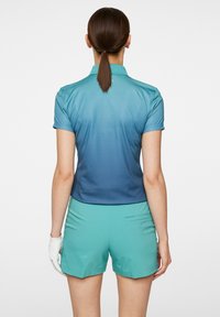 Poloshirt met korte mouwen in een ombre-design, van licht naar donkerblauw, gecombineerd met teal korte broek. Materiaal lijkt licht en soepel te zijn.