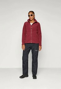 Μαραντί φλις τζάκετ με ψηλό γιακά και κλείσιμο με φερμουάρ. Μαύρο αδιάβροχο παντελόνι, λεπτομέρεια λογότυπου και μαύρα υποδήματα. Μοντέλο που φοράει γυαλιά ηλίου.