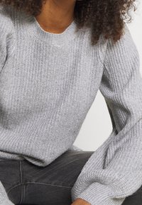 Grå stickad tröja med ribbad textur, rund halsringning och breda, utställda ärmar. Tyget verkar mjukt och varmt.