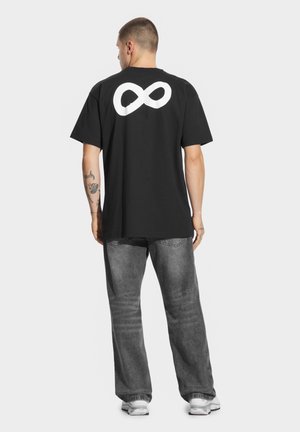 Czarny bawełniany T-shirt z dużym białym symbolem nieskończoności na plecach, w połączeniu z szarymi dżinsami i białymi trampkami. Prosty design z przodu.