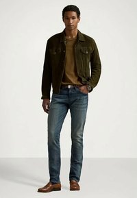 Casaco de camurça verde escuro sobre uma camisa bege, combinado com jeans de ganga azul e sapatos de couro castanhos. Relógio no pulso completa o visual.