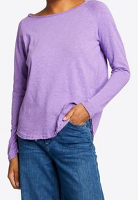 Langärmliges lila Oberteil aus einem weichen, leichten Stoff mit lockerer Passform, Rundhalsausschnitt und rohem Saum. Kombiniert mit blauen Jeans.