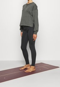 Šedá mikina s kleslými rameny a uvolněným střihem kombinovaná s černými, přiléhavými legínami. Naboso na jóga podložce.