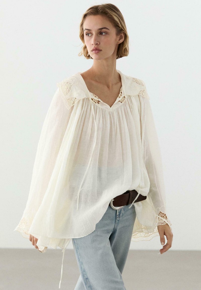 Donna con una camicetta bianca ampia e trasparente con dettagli in pizzo e jeans azzurri, in piedi davanti a uno sfondo chiaro e uniforme.