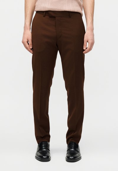 Άνδρας που φορά καφέ ραμμένο παντελόνι και μαύρα δερμάτινα επίσημα παπούτσια, στέκεται με χαλαρά τα χέρια δίπλα στο σώμα.