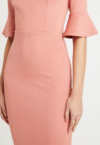 Sydd rosa klänning med ärmar som går till armbågen och klockformade, tillverkad av ett mjukt tyg. Har en figurnära silhuett och minimal detaljering.