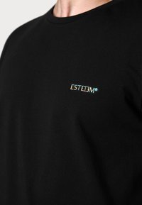 Schwarzes Baumwoll-T-Shirt mit einem kleinen, bunten "ESTEEM"-Logo auf der Brust. Der Stoff hat eine glatte, leicht elastische Textur.