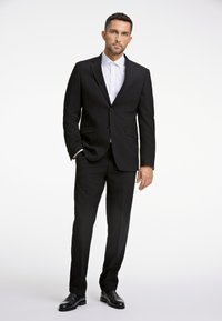 Zwart op maat gemaakt pak met een jacket met één knoop, wit overhemd met kraag en zwarte leren schoenen. Soepele stof, moderne pasvorm en strakke lijnen.