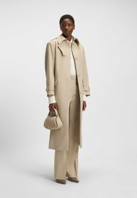 Beige tailored trench coat with a collar and gold buttons, paired with wide-leg trousers and a small beige handbag, set against a neutral background.
