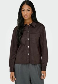 Chaqueta de camisa de ante marrón con cuello, botones de presión, dos bolsillos en el pecho y mangas largas, mostrada con pantalones grises.