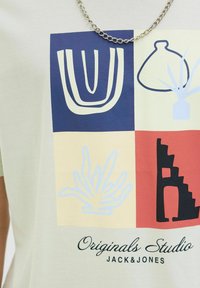 Lysfarvet T-shirt med grafisk print i fire firkanter: abstrakte former, planter og en sort struktur. Tekst nederst.