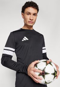 Zwart sportshirt met lange mouwen en witte strepen op de mouwen, met het Adidas-logo; houdt een met patronen versierde zwart-witte voetbals. 