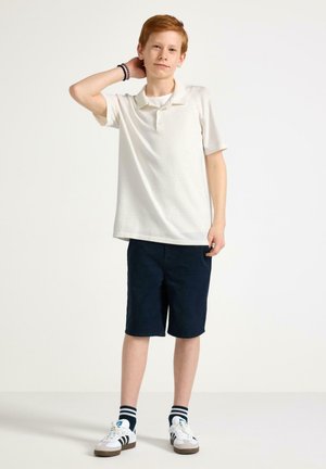 Rødharret dreng iført hvid polo, mørke shorts, stribede sokker og hvide sneakers står med den ene hånd bag hovedet mod en ensfarvet baggrund.