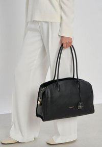 Personne en tenue blanche tenant un sac à main en cuir noir Lancaster Paris avec double poignées et détail de fermeture éclair dorée.