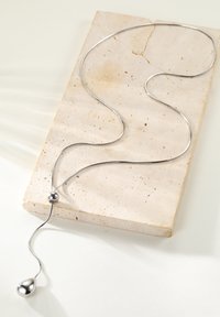 Zilveren halsketting met een gladde, langwerpige schakel en ronde kraalaccenten; tentoongesteld op een gestructureerd, lichtgekleurde stenen oppervlak.