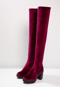 Des bottes en velours magenta profond, hautes, avec des talons carrés et des bouts ronds, se tenant droites sur une surface blanche contre un fond uni.