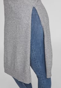 Ljust grått stickat tröja med ribbat avslut och sidofickor, bärs över mörkblå jeans, med en mjuk textur.