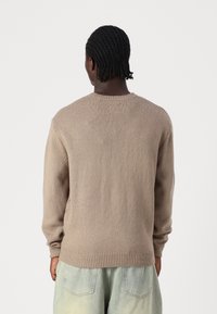 Beige gestrickter Pullover mit Rundhalsausschnitt, gerippten Bündchen und Saum, mit einer strukturierten Oberfläche und langen Ärmeln, getragen über hellen Hosen.