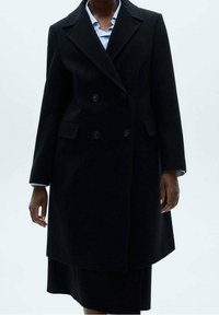 Manteau noir croisé avec revers crantés, poches latérales et coupe ajustée. Fabriqué en tissu de laine texturé. Manches longues.