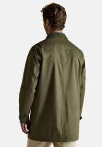 Vīrietis, kurš valkā garu olīvu zaļu mētelī ar korduroja apkakli, stāv ar muguru pret skatītājam uz vienkārša fona.