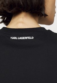 Černé tričko s kulatým výstřihem a bílým logem "KARL LAGERFELD" na horní části zad. Měkká bavlněná tkanina, minimalistický design.