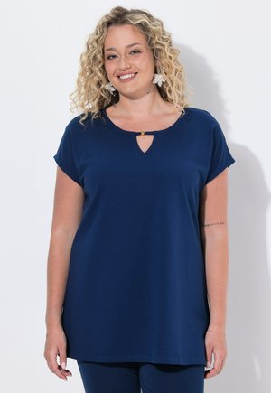Lächelnde Frau mit lockigem blonden Haar trägt ein marineblaues kurzärmliges Oberteil mit kleinem Schlitz und goldenem Akzent am Ausschnitt.
