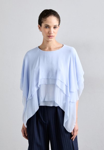 Haut superposé bleu clair avec des drapés en tissu fluide, texture douce, et un décolleté arrondi. Assorti à un pantalon sombre à rayures fines.