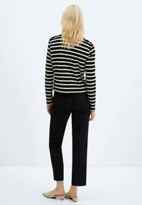 Donna con i capelli biondi lunghi che indossa una camicia a righe bianca e nera a maniche lunghe, pantaloni neri e scarpe beige a tacco basso, in piedi di spalle.