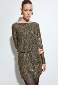 Robe en sequins dorés scintillants avec des manches longues, un col bateau et une taille froncée, réfléchissant la lumière grâce à une surface texturée.