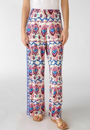 Frau trägt weite Hosen mit bunten geometrischen und abstrakten Mustern, kombiniert mit beigen Sandalen und einem weißen Oberteil.